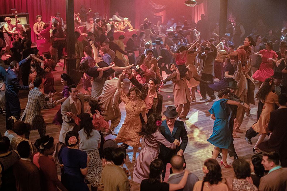 A crowd of dancers dressed in suits and dresses suitable for 1930s and '40s America improvise on the dance floor, a brass band just visible along the back wall.